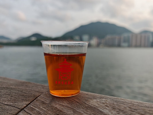 A Pu-erh tea set on a white ceramic plate, featuring scattered dark-brown Pu-erh tea leaves, a glass cup filled with amber tea liquor, and a vintage brown ceramic canister containing tea leaves. Background is a blurred green lawn.