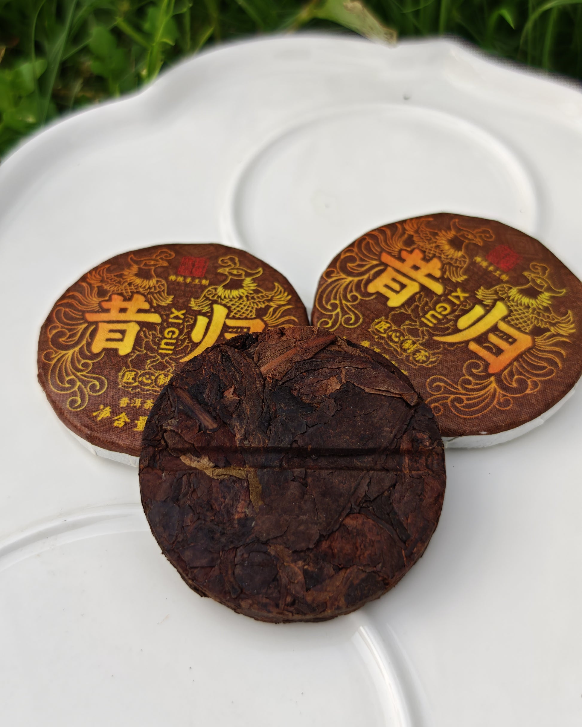 白色瓷盤上的昔歸普洱熟茶展示。包含一小堆散茶葉、一塊圓形的普洱茶餅，以及一個用竹編外殼和繩子包裝的迷你茶筒，標籤上印有「昔歸（熟茶）」繁體字樣。背景為模糊的綠色草地。昔歸普洱熟茶-茶餅與竹編包裝展現