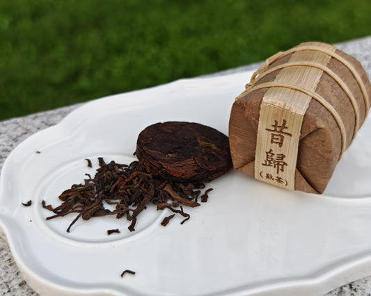 白色瓷盤上的昔歸普洱熟茶展示。包含一小堆散茶葉、一塊圓形的普洱茶餅，以及一個用竹編外殼和繩子包裝的迷你茶筒，標籤上印有「昔歸（熟茶）」繁體字樣。背景為模糊的綠色草地。昔歸普洱熟茶-茶餅與竹編包裝展現